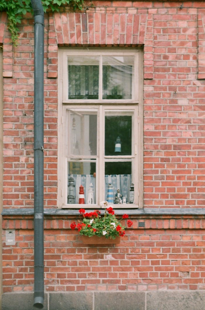 about-01 A rustic brick wall featuring a window with vibrant red flowers in a quaint urban setting.